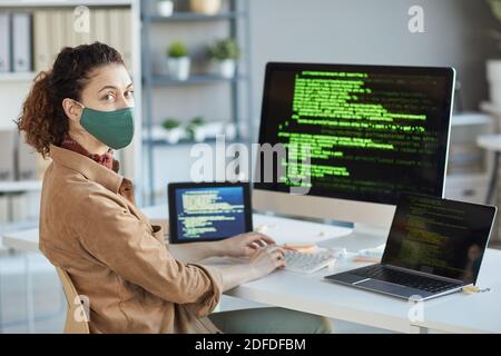 Porträt des jungen Programmierers in Schutzmaske Blick auf die Kamera Während sie an ihrem Arbeitsplatz mit Computern auf dem Tisch sitzt Stockfoto