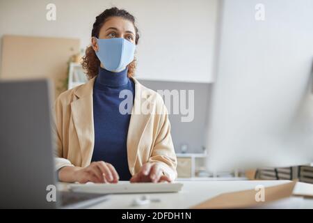 Junge Frau in Schutzmaske Blick auf Computer-Monitor sie Online-Arbeit am Tisch während der Pandemie Stockfoto