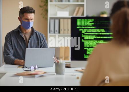 Reife Geschäftsmann in Schutzmaske arbeiten auf Laptop an der Gemeinsam mit seinem Kollegen, der im IT-Büro arbeitet, an einem Tisch Stockfoto