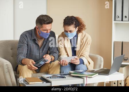 Zwei Designer in Masken sitzen auf dem Sofa und untersuchen die Fotos während der Teamarbeit im Büro Stockfoto