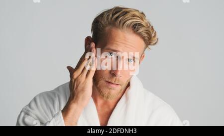 Attraktive bärtige Mann im Bademantel Anwendung Creme auf Gesicht isoliert Auf weißem Hintergrund Stockfoto