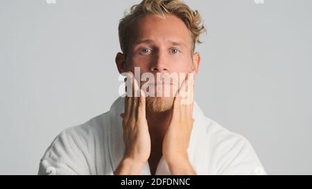 Hübscher bärtiger Mann im Bademantel, der intensiv Creme auf das Gesicht aufträgt Blick in die Kamera auf weißem Hintergrund Stockfoto