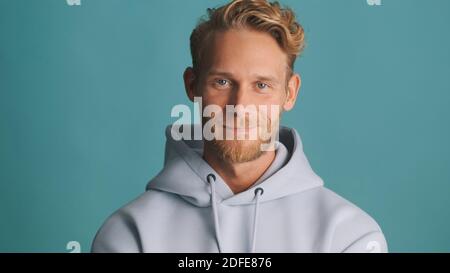 Attraktive stilvolle bärtige Kerl in Kapuzenpullover intenly suchen in der Kamera Und lächelnd isoliert auf blauem Hintergrund Stockfoto