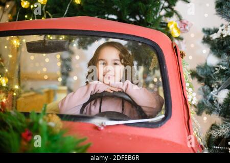 Kleine Vorschulmädchen sitzen hinter Lenkrad in Dekor Papier Rotes Auto auf magischem Hintergrund mit Weihnachten und Neujahr Tannenbaum und Lichter Stockfoto