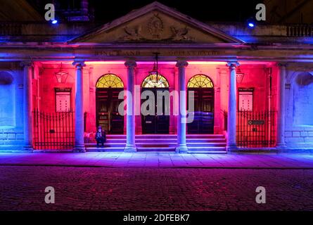 Bath, Großbritannien. Dezember 2020. Eine Frau, die in Licht gebadet wird, sitzt auf den Stufen der Pump Rooms, während sie an einem klaren Winterabend auf ihr Handy schaut.um die dunklen Tage aufzuhellen, hat Bath BID einige spektakuläre Denkmalbeleuchtung in einigen der ikonischen Straßen von Bath installiert.die Installationen haben Entworfen und produziert von Fineline Lighting and Visit Bath und die Straßen werden jeden Tag bis zum Ende des Jahres beleuchtet. Quelle: Lynchpics/Alamy Live News Stockfoto