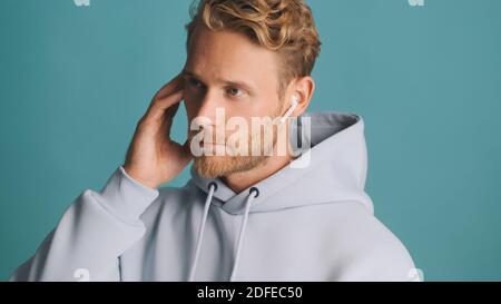 Gut aussehende stilvolle bärtige Kerl in Kapuzenpullover suchen ernsthafte Gespräche auf drahtlosen Kopfhörer isoliert auf blauem Hintergrund. Moderne Technik Stockfoto