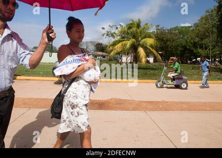 Junge Frau trägt ihr Baby mit Mann hält einen Sonnenschirm, um sie vor der Sonne und Menschen auf einem Rollstuhl im Hintergrund zu schützen. Puerto Lempira, Mosquitia, Honduras Stockfoto