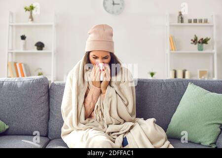 Junge kranke Frau in warmer Kleidung, Hut auf Sofa unter Decke sitzend, Nase bläst und krank fühlen Stockfoto