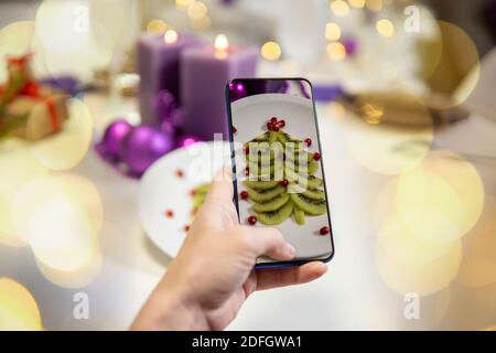 Mädchen Hand macht Bilder von Weihnachtstisch. Kiwi und Granatapfel in Form eines weihnachtsbaums auf einem Teller. Hohe Qualität Stockfoto