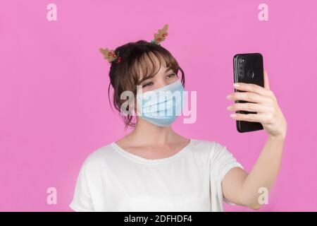 Frau trägt schützende Gesichtsmaske und Videoanruf für Weihnachten. Neues Konzept für die Feier. Rosa Hintergrund. Hochwertige Fotos Stockfoto