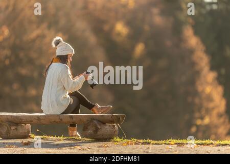 Fotografin in warmen Kleidern macht Bilder von der Natur im Freien, sitzt auf einer Holzbank. Stockfoto