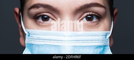 Junge kaukasische Frau trägt blaue chirurgische Gesichtsmaske, Nahaufnahme Widescreen-Porträt, Unsicherheit In Augen, isoliert auf dunkelgrauem Hintergrund, Coronavirus COV Stockfoto