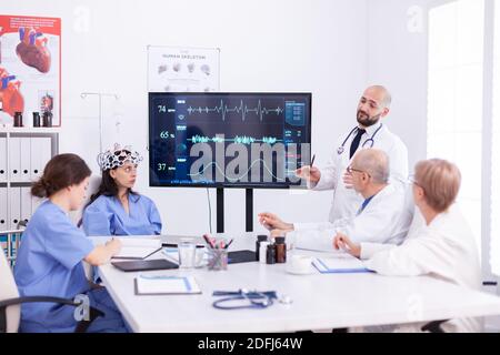 Arzt diskutiert über die Zukunft der Neurologie mit medizinischem Personal im Besprechungsraum des Krankenhauses. Der Monitor zeigt moderne Gehirnstudien, während ein Wissenschaftlerteam das Gerät anpasst. Stockfoto