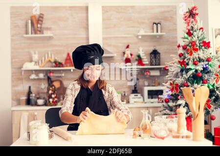 Fröhliches Kind spielen mit Teig am weihnachtstag trägt Schürze.Fröhlich glücklich niedlichen Mädchen beim Vorbereiten leckere Cookies für weihnachtsfeier in der Küche mit weihnachtsbaum im Hintergrund. Stockfoto