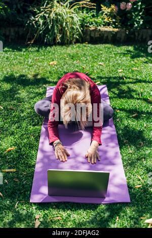 Online Sport Fitness Yoga Training Workout, Senior Frau im Garten ihres Hauses macht Übungen auf Pilates Matte gegenüber Laptop zu Hause. Stockfoto
