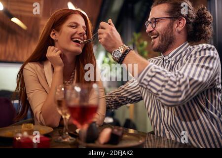 Ein junger Mann füttert seine Freundin am Valentinstag Feier in einem Restaurant in einer fröhlichen Atmosphäre. Gemeinsam, Jubiläum, Feier Stockfoto