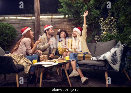 Multiethnische Gruppe von Freunden feiern Silvester, singen, lächeln, lachen, mit santa Klausel Hüte auf den Köpfen und Gläser Weißwein i Stockfoto