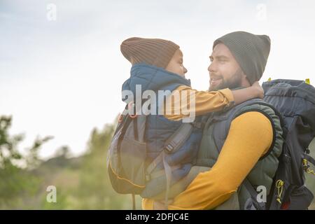 Junge liebevolle Vater mit Rucksack hält seinen niedlichen kleinen Sohn auf den Händen gegen natürliche Umgebung gegen Himmel, grünes Gras und Sträucher Stockfoto