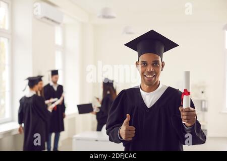 Dunkelhäutige afroamerikanische Absolventin mit Diplom und hebt den Daumen hoch und schaut auf die Kamera. Stockfoto