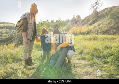 Junges Paar und ihre niedlichen kleinen Sohn mit Rucksäcken stehen Auf dem Boden vor der Kamera, während der Mann zu gehen Teppich ausrollen, um Ruhe zu haben Stockfoto