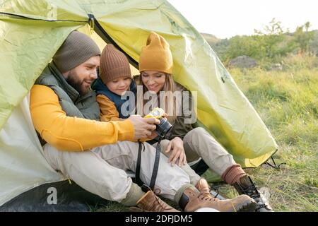 Junger bärtiger Mann, der seiner Familie Fotos in Fotokamera zeigt Während der Erholung im Zelt in natürlicher Umgebung im Herbst Tag während der Reise Stockfoto