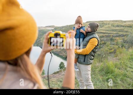 Junge Frau mit Fotokamera fotografieren ihren Mann und Kleiner Sohn steht gegen See umgeben von Bergen während der Reise Stockfoto