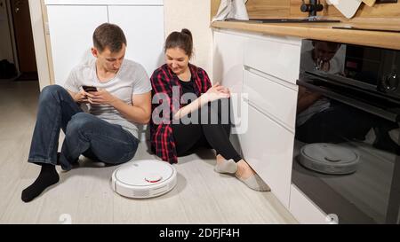 Glückliche junge Familie sitzt auf dem Boden Steuerung zeitgenössischen Roboter Vakuum Reiniger Entfernen von Schmutz vom Holzboden mit Smartphone in der Küche Stockfoto