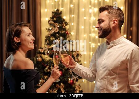 Seitenansicht eines jungen fröhlichen Paares, das sich anschaut Mit einem Lächeln, während vor dem Zimmer mit Sektflöten klirren Der Kamera von Weihnachtsbaum Stockfoto