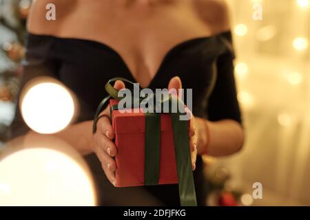 Hände von jungen eleganten weiblichen in schwarzen Abendkleid halten Rot verpackte geschenkbox gebunden mit dunkelgrünem Band im Stehen Vor der Kamera Stockfoto