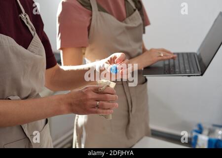 Hand der Frau in Schürze setzen handgemachte blaue Glasperlen In kleinen Leinen- oder Baumwollsack, während ihr Kollege mit Laptop, der Daten eingibt Stockfoto