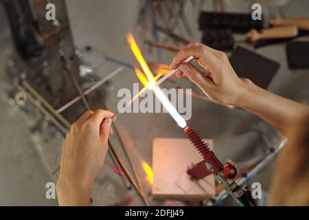 Hände von jungen Handwerker sitzen am Arbeitsplatz im Studio und Brennen von zwei stäbchen geformten Werkstücken im Brenner und Halten In Brand Stockfoto