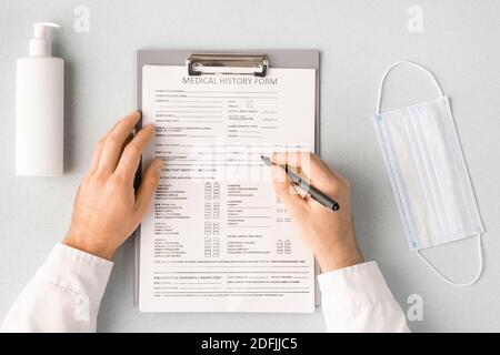 Übersicht über die Hände des Arztes mit Stift über die Krankengeschichte Formular ausfüllen der persönlichen Daten des Patienten mit Maske und Desinfektionsmittel in der Nähe Stockfoto