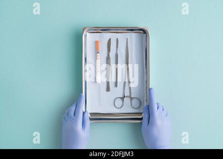 Blick auf Handschuhen des Chirurgen setzen Stahlschale mit Chirurgische Instrumente und Spritze auf staubblauem Hintergrund vor der Operation Stockfoto