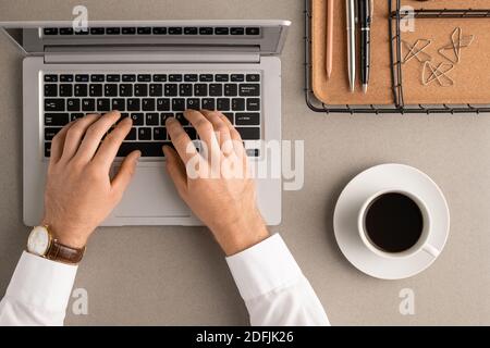 Draufsicht auf die Hände des zeitgenössischen Managers, der die Tasten drückt Laptop-Tastatur während der Vernetzung zwischen Bürobedarf und Tasse Kaffee Stockfoto