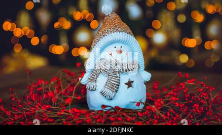 Schöne Weihnachten Home Dekoration. Niedliches kleines Schneemann-Spielzeug, das im roten Beerenkranz auf dem Tisch über glühenden Weihnachtsbaum Bokeh-Lichtern steht Stockfoto