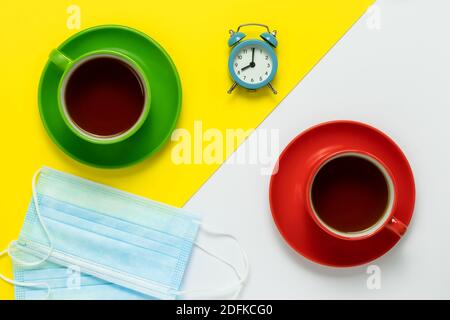 Teetrinken. Im Café befinden sich zwei blaue medizinische Schutzmasken auf dem Tisch. Zwei Tassen aus Keramik mit roten Untertassen und Kaffee auf einem gelben und weißen Stockfoto