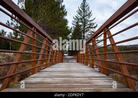 Ein Blick von der Mitte einer hölzernen Fußgängerbrücke über einen kleinen Bach. Rostiger, verwitternder Stahl bildet Geländer, die sich über beide Seiten erstrecken. Leichter Schnee, ev Stockfoto