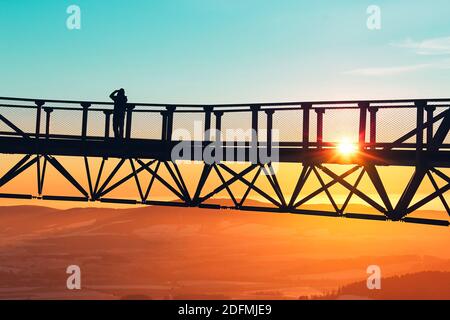 Silhouette des Menschen auf der Brücke des Aussichtsturms Stezka V oblacích in Tschechien mit Blick auf schneebedeckte Hügel und Bäume bei farbenfrohem Sonnenuntergang. Stockfoto