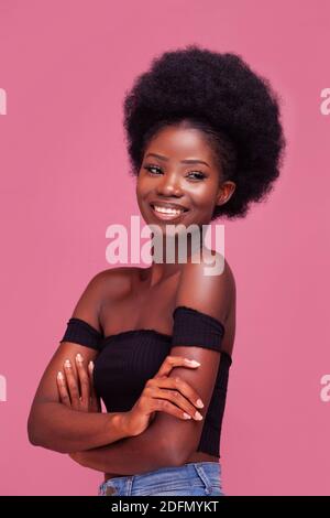 Happy Charming African American Mädchen mit erstaunlichen afro Haare stehen Lächelnd mit Arm gefaltet in schwarzem Top und Jeans Isoliert auf rosa Hintergrund Stockfoto