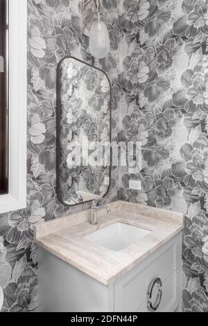 Ein modernes Badezimmer mit gemusterter Tapete, grauem Schminktisch und Marmorablage sowie einer schönen Leuchte über dem bronzenen Spiegel. Stockfoto