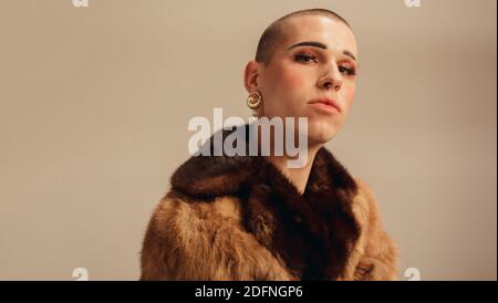 Porträt eines hübschen jungen Mannes im Pelzmantel. Geschlecht Flüssigkeit männlich mit Make-up und Ohrring vor beigem Hintergrund. Stockfoto