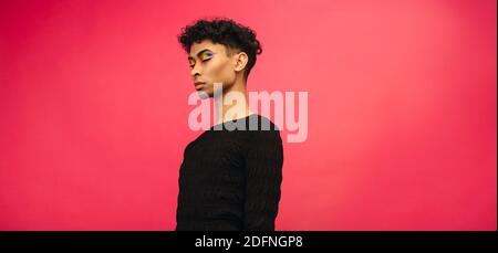 Junger Mann mit Regenbogen Augenschatten Make-up auf rotem Hintergrund mit geschlossenen Augen stehen. Geschlecht fließender Mann in schwarzem T-Shirt. Stockfoto