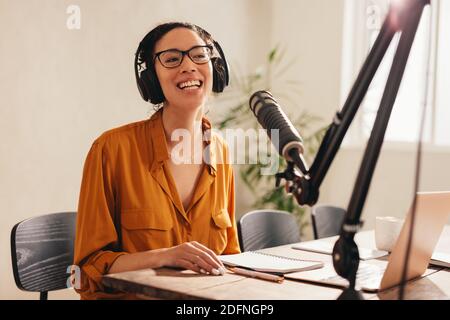 Frau mit Kopfhörer am Tisch mit Laptop und Mikrofon sitzen. Frau, die die Kamera anschaut und lächelt, während sie einen Podcast von zu Hause aufzeichnet. Stockfoto