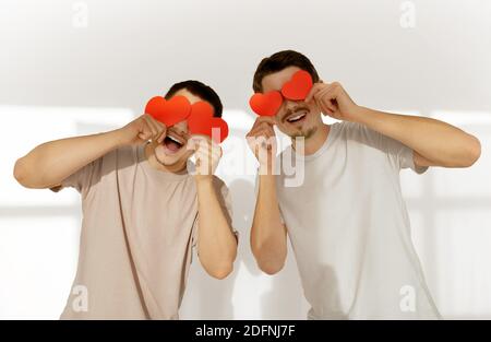 Glückliches Paar von jungen Homosexuell Männer verstecken ihre Augen hinter Rotes Papier valentine Herzen zur Feier zu Hause gegen weiß Wandhintergrund Stockfoto