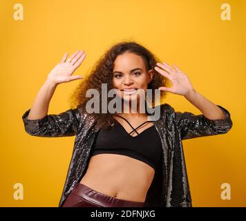 Mode schöne afroamerikanische Modell junge Frau mit langen lockig Haare posieren tanzen auf der Kamera mit Händen oben tragen glänzend Jacke und schwarzes Shirt Stockfoto