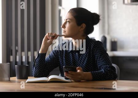 Junge hindu-Frau, die am Schreibtisch studiert, schaut beiseite und hält Telefon Stockfoto