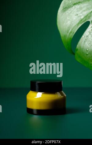 Glas Glas mit natürlicher Feuchtigkeitscreme und tropischem Blatt auf grünem Hintergrund. Verpackungsdesign für Bio-Kosmetik. Stockfoto