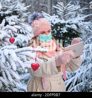 Glückliche Frau im Gesicht Maske macht Selfie Foto auf Tablet im Winter Park in der Nähe des Weihnachtsbaumes - Moskau, Russland, 02. Dezember 2020 Stockfoto