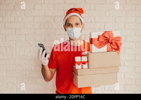 Junger Mann Kurier in medizinischen Gesichtsmaske, Schutzhandschuhe, weihnachtsmütze liefern Weihnachtsgeschenke. Glückliche Lieferung Mann im Gesicht Maske halten Smartphone, Geschenk, Stockfoto