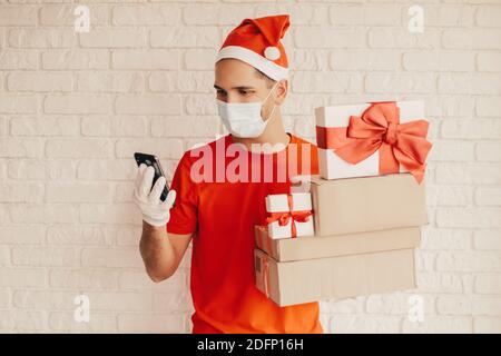 Junger Mann Kurier in medizinischen Gesichtsmaske, Schutzhandschuhe, santa hat verwenden mobile Lieferung App auf Smartphone. Glückliche Lieferung Mann in Gesichtsmaske tragen gif Stockfoto